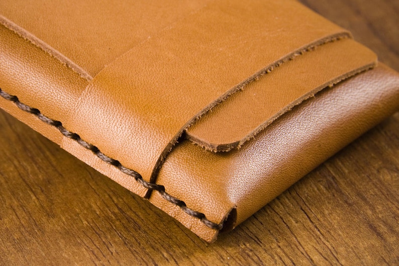 Puede incluir: Primer plano de una cartera de cuero marr&oacute;n con un borde cosido. La cartera est&aacute; doblada por la mitad y tiene una superficie lisa y texturizada.