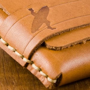 May include: Close-up of a brown leather wallet with a stitched edge and a silhouette of a surfer on the front.