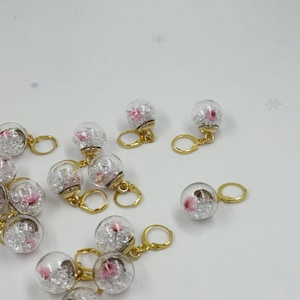Puede incluir: Pendientes de bola de cristal transparente con pequeñas flores rosas y aros dorados. Los pendientes tienen un diseño floral delicado, con un interior brillante en forma de cristal. Fondo blanco.
