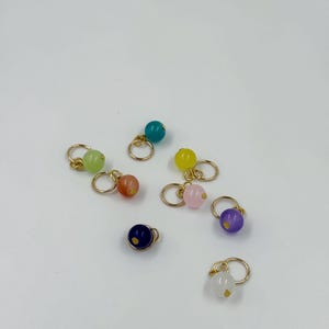 Peut inclure: Une collection de boucles d'oreilles à perles colorées. Chaque boucle d'oreille présente une petite perle ronde de couleur différente, notamment verte, orange, jaune, rose, violette, bleue et blanche. Les perles sont fixées à de petits cerceaux dorés.
