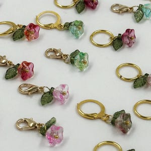 May include: A collection of floral charms with gold-coloured clasps and hoops. The charms feature translucent flowers in pink, green, and blue, with small green leaves. These charms are suitable for jewellery making or craft projects.