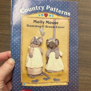 May include: A book cover titled "Country Patterns" featuring "Molly Mouse Doorstop & Broom Cover." The cover has a blue background with white dots and an illustration of two mouse figures wearing aprons. The text "Ozark Crafts Inc." is also visible.