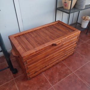 Pode incluir: Um baú de armazenamento retangular de madeira com acabamento em castanho quente, apresentando um design de ripas na tampa e nas laterais. O baú possui uma tampa articulada com um pequeno puxador de madeira centrado na borda frontal.