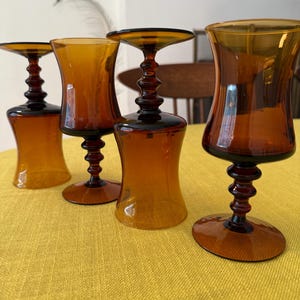 May include: Four amber-colored stemmed glasses with a decorative, ridged stem and a flared bowl. The glasses are arranged on a yellow tablecloth, showcasing their vintage design and warm tones. The glasses are likely for serving beverages.