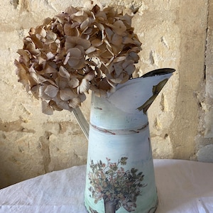 May include: A white metal pitcher with a floral design and a dried hydrangea bouquet. The pitcher has a rustic, vintage look and is perfect for adding a touch of farmhouse charm to your home decor.