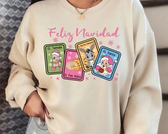 May include: A beige sweatshirt with the text "Feliz Navidad" in pink lettering. The sweatshirt features a design of four colorful cards with cartoon characters and the text "El Tamale", "El Burrito Sabanero", "El Pan Dulce", and "El Santa".