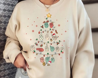 May include: A beige sweatshirt with a Christmas tree design featuring hand signs for the word "Merry" in American Sign Language. The tree is decorated with ornaments, lights, and a star on top.