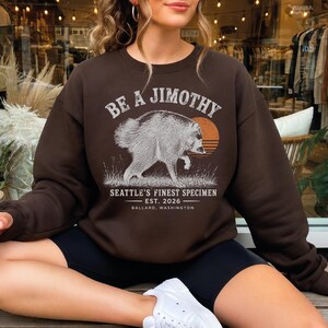 A woman wearing a brown sweatshirt with a graphic design of a jaguar and text, sitting on a bench and holding a drink.