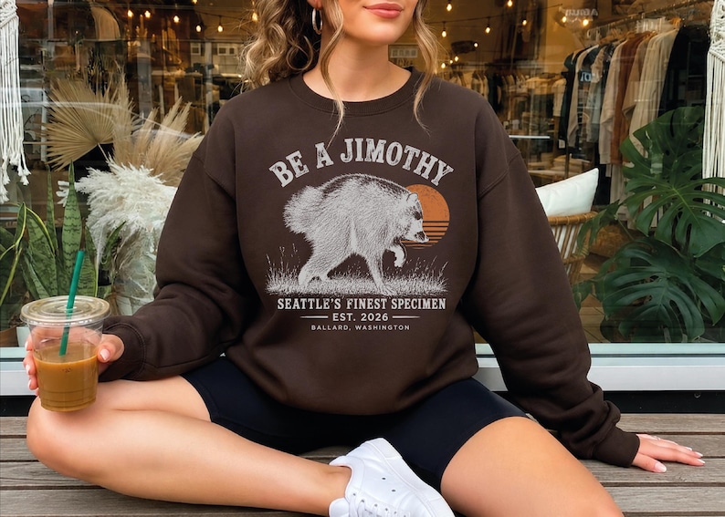A woman wearing a brown sweatshirt with a graphic design of a jaguar and text, sitting on a bench and holding a drink.