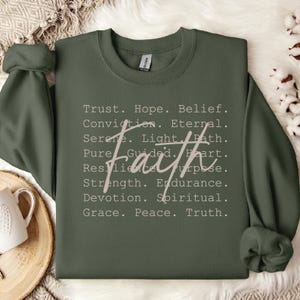 Peut inclure: Un sweat-shirt à col rond vert olive avec une liste de mots incluant Trust, Hope, Belief, Conviction, Eternal, Serene, Light, Path, Pure, Guided, Heart, Resilience, Purpose, Strength, Endurance, Devotion, Spiritual, Grace, Peace, Truth, et le mot faith écrit en grande écriture cursive au centre.