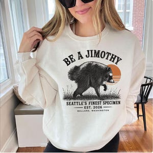 A woman wearing a white sweatshirt with a graphic design of a bear and text that says "Be a Jimothy Seattle's Finest Specimen Est. 2026 Ballard, Washington".