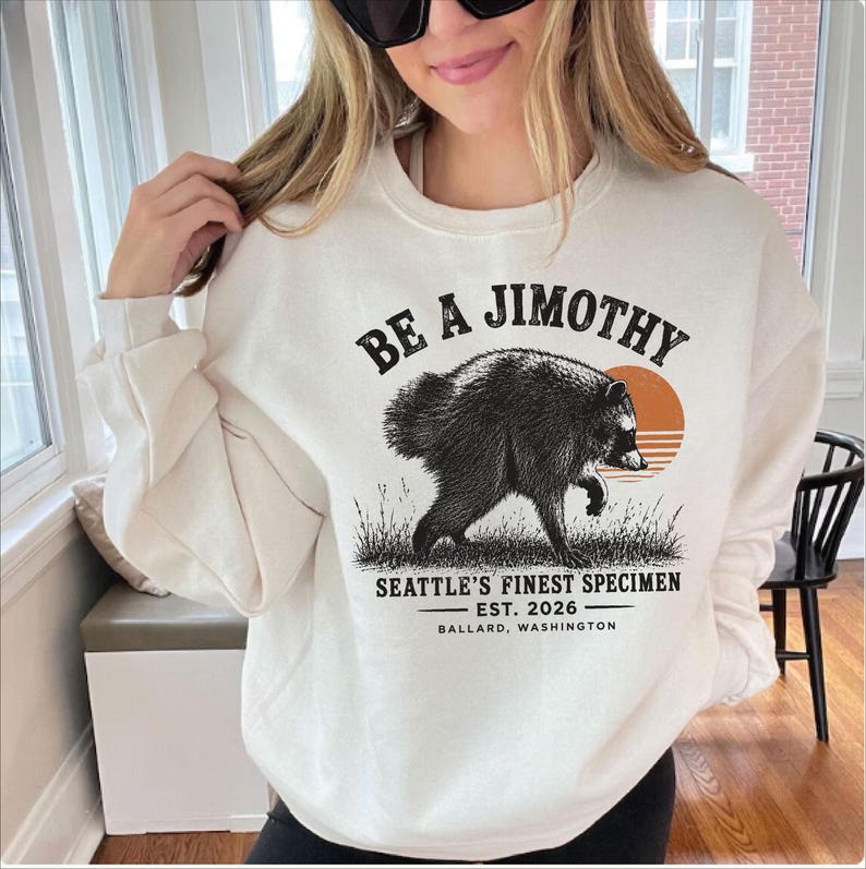 A woman wearing a white sweatshirt with a graphic design of a bear and text that says "Be a Jimothy Seattle's Finest Specimen Est. 2026 Ballard, Washington".