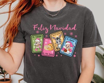 May include: A dark gray t-shirt with the text "Feliz Navidad" in pink letters. The shirt features a design of five colorful cards with illustrations of traditional Mexican Christmas foods and characters. The cards are labeled "El Tamale", "El Burrito Sabanero", "El Pan Dulce", and "El Santa".