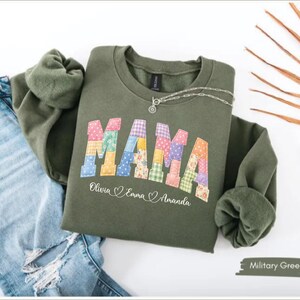 May include: An olive green sweatshirt with the word "MAMA" in colorful patchwork letters. Below the word are the names Olivia, Emma, and Amanda. A silver necklace rests on the sweatshirt, which is paired with blue jeans.