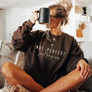 May include: A dark brown sweatshirt with the text "NOT PERFECT just forgiven" in white. A black mug is held up to the face. The sweatshirt has a cross graphic above the word "PERFECT".