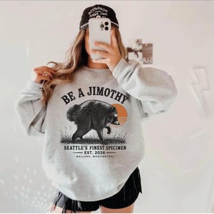 A person is taking a selfie with a grey sweatshirt featuring a graphic of a bear and text that says "Be a Jimothy Seattle's Finest Specimen Est. 2026 Balland, Washington".
