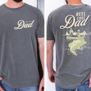 May include: Gray t-shirt with "Reel Cool Dad" text on the front and a fishing graphic on the back. The graphic features a bass, a canoe, and two people fishing. The shirt is a crew neck.