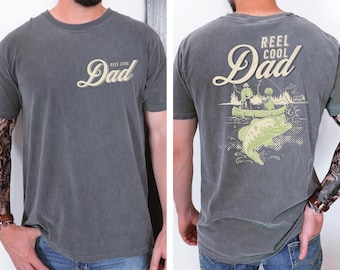 Pode incluir: Camiseta cinza com o texto "Reel Cool Dad" na frente e um gráfico de pesca nas costas. O gráfico apresenta um baixo, uma canoa e duas pessoas pescando. A camisa é de gola redonda.
