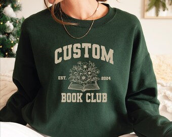 May include: Dark green sweatshirt with the text "CUSTOM BOOK CLUB" in a light beige color. The design includes an open book with flowers and the year "2024". The sweatshirt is a casual garment.