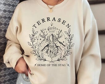 May include: A beige sweatshirt with a black graphic design featuring a stag, mountains, and stars. The text "TERRASEN" and "HOME OF THE STAG" are printed on the sweatshirt.