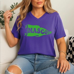 May include: A purple v-neck t-shirt with a green pennant design that reads "MASCOT" in a playful font. The shirt is made of a soft material and is paired with blue jeans. The overall style is casual and comfortable.