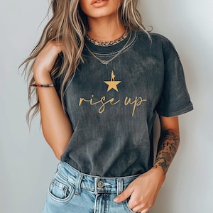 Peut inclure: T-shirt gris foncé avec l'inscription "rise up" en lettres dorées sous une étoile et une silhouette dorées. Le t-shirt a un col rond et des manches courtes. Le t-shirt est porté avec un jean bleu.