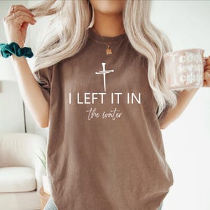 A woman with long blonde hair wearing a brown t-shirt with the text "I LEFT IT IN the winter" printed on it. She is holding a coffee mug in her right hand.