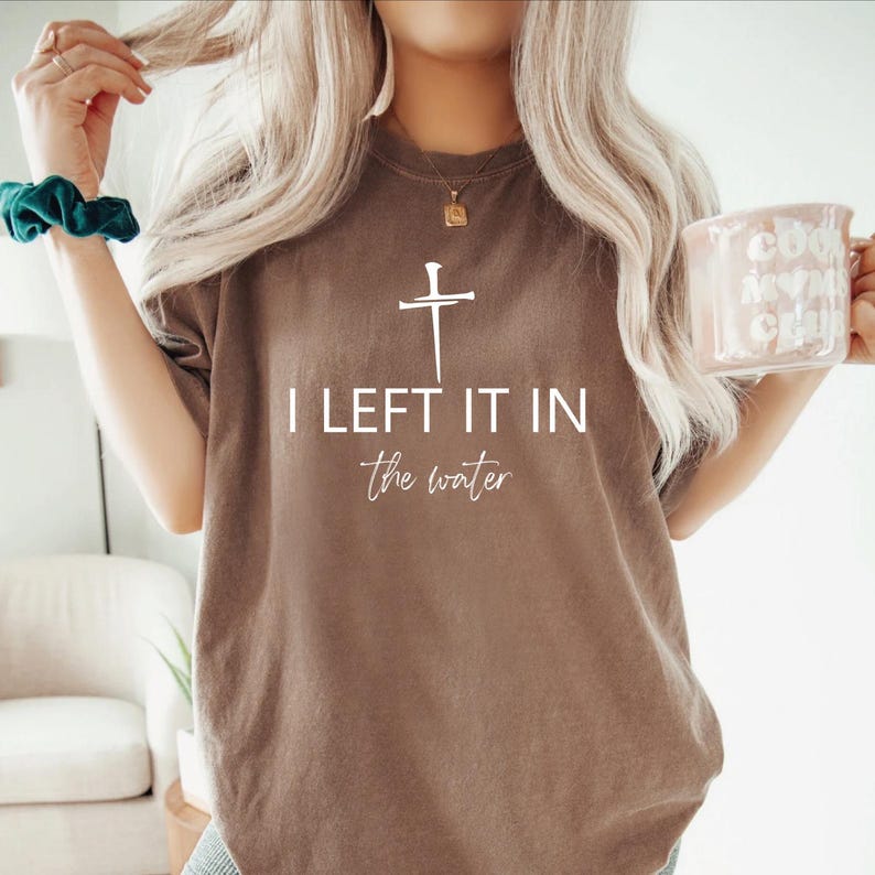 A woman with long blonde hair wearing a brown t-shirt with the text "I LEFT IT IN the winter" printed on it. She is holding a coffee mug in her right hand.