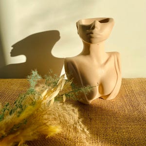 Op de afbeelding: Een beige keramische plantenpot in de vorm van een vrouwenbuste met een uitgeholde bovenkant om te planten. De pot staat op een bruine jute ondergrond met gedroogde bloemen.