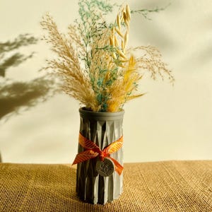 Könnte beinhalten: Eine kleine graue Betonvase mit strukturierter Oberfläche, verziert mit einem orangefarbenen Band und einem kleinen Etikett. Die Vase ist mit getrockneten Gräsern und Blumen in Gelb- und Grüntönen gefüllt.