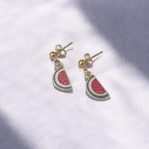 K&ouml;nnte beinhalten: Ein Paar goldene Wassermelonen-Scheiben-Ohrringe. Die Ohrringe haben eine rote Wassermelonen-Scheibe mit gr&uuml;ner Schale und wei&szlig;en Kernen.