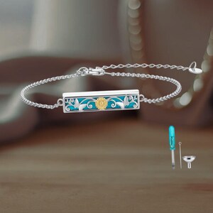 May include: Silver bracelet with a rectangular pendant featuring a turquoise background, gold sunflower, and butterfly accents. Includes a small blue tool, a silver tool, and a small silver funnel.
