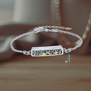 Puede incluir: Pulsera de plata con un colgante rectangular. El colgante tiene una incrustación azul-verde y está decorado con flores y hojas doradas. La pulsera tiene un diseño de cadena delicado y un cierre seguro.