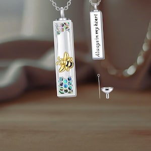 May include: Silver-toned rectangular pendant necklace with a bee design. The pendant features a gold-colored bee and iridescent honeycomb-shaped accents. A second pendant reads "Always in my heart". Includes a small funnel and tool.