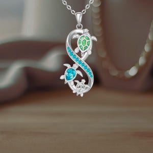 May include: Silver pendant necklace with three turtle charms. The pendant has a flowing design with blue and green gemstone accents. The turtles have turquoise and green gemstone shells. The necklace is suspended from a delicate chain.