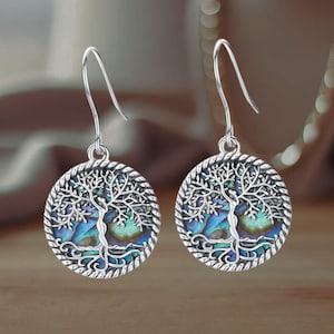 May include: A pair of silver earrings featuring a tree of life design. The tree is crafted from abalone shell and is encircled by a silver rope border.