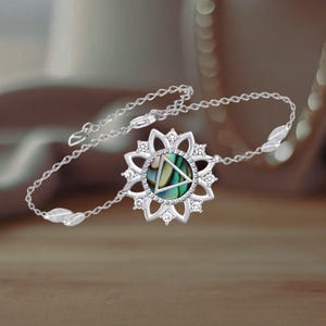 May include: A silver bracelet with a delicate chain and a circular pendant featuring a green and blue abalone shell with a triangle design. The pendant is surrounded by a floral design with clear crystals.