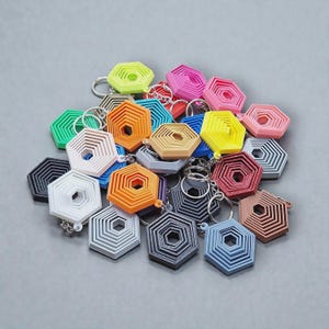 May include: A collection of colorful, geometric keychains in various shades including green, pink, orange, and blue. Each keychain features a layered hexagonal design with a silver keyring attached. The keychains are arranged on a gray surface.
