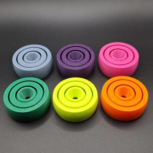 May include: Six colorful, concentric ring-shaped objects in shades of blue, purple, pink, green, yellow, and orange. Each object has a cylindrical center and a smooth, matte finish. The objects are arranged in two rows of three against a dark background.