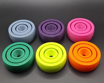 May include: Six colorful, concentric ring-shaped objects in shades of blue, purple, pink, green, yellow, and orange. Each object has a cylindrical center and a smooth, matte finish. The objects are arranged in two rows of three against a dark background.