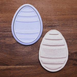 May include: Two egg-shaped cookie cutters. One is light purple and the other is beige. Both have a similar design with horizontal lines and a rectangular section in the middle. The cutters are on a wooden surface.