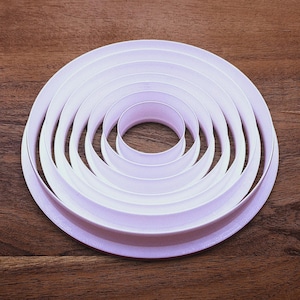 May include: A set of six concentric, circular cookie cutters in varying sizes. The cutters are made of white plastic and are arranged in a nested pattern on a wooden surface. The largest cutter has a diameter of approximately 6 inches.