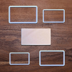 May include: Rectangular cookie cutters in various sizes, along with a rectangular piece of dough, are arranged on a wooden surface. The cutters are white, and the dough is a light pink color. The arrangement suggests a baking or crafting theme.