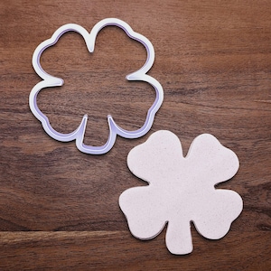 May include: A clover-shaped cookie cutter and a cut-out cookie are displayed on a wooden surface. The cookie cutter is white with a lavender interior, and the cookie is light pink with a speckled texture. The items are for baking.