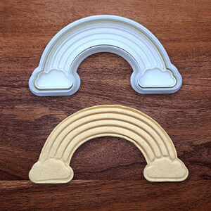May include: Two rainbow-shaped cookie cutters. One is white and the other is beige. Each cutter has a rainbow shape with clouds on each end. The cutters are made of plastic and are designed for baking.