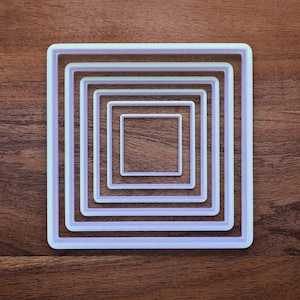 May include: A set of five nested square cookie cutters in varying sizes. The cutters are white with a light purple hue and are arranged on a dark brown wooden surface. The square shapes are evenly spaced, creating a layered effect.