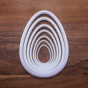 May include: A set of white plastic egg-shaped cookie cutters of varying sizes, nested together. The cutters are arranged on a dark brown wooden surface, creating a layered effect. The shapes range from large to small, ideal for baking.