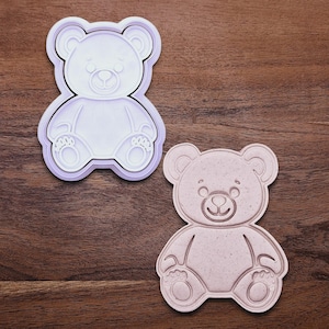 May include: Two teddy bear-shaped cookie cutters. One is white with a purple outline, and the other is a light pink color. The cutters have detailed features, including the bear's face, paws, and body. The cutters are on a wooden surface.
