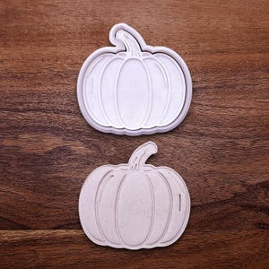 May include: Two pumpkin-shaped cookie cutters. One is white with a detailed interior design, and the other is a light beige color. Both cutters have a stem and vertical lines to represent the pumpkin's segments. The cutters are on a wooden surface.