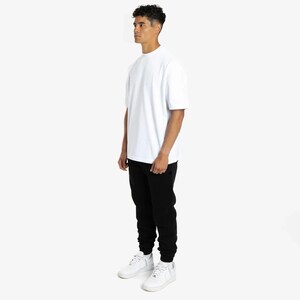 Puede incluir: Una persona que lleva una camiseta blanca de cuello redondo, pantalones de ch&aacute;ndal negros y zapatillas blancas. La camiseta tiene mangas cortas. Los pantalones de ch&aacute;ndal tienen cintura con cord&oacute;n y tobillos con pu&ntilde;os. Las zapatillas son bajas y con cordones.